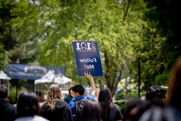 A 博彩网址大全 101 leader seen from behind in a crowd gives a tour to new students holding a sign that says Follow Me